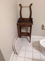 Wood wash stand with drawer and bottom shelf, two beige chair pads leaning against the wall beside it, set in white tiled bathroom.