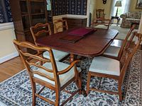 Full view of wooden dining table with leaf inserted, six chairs around table, protective covers on tabletop, placed on patterned rug in dining room.
