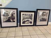 Three framed black and white prints lined up on floor against wall, displaying bridge and canal scenes.