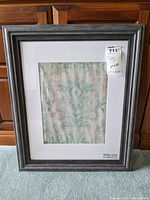 Framed wall art print standing upright against a wooden furniture, visible gray wooden frame and white mat with a decorative print inside.