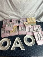 Photo showing multiple metallic cake topper letters in pink and white packaging and three large white ceramic letter dishes O, A, O.
