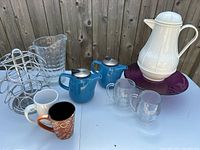 Photo of coffee and tea set including 2 blue kettles, measuring pitcher, 2 glass mugs, 2 ceramic mugs, a white thermos, and purple cake plate