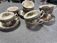Set of eight teacups stacked and arranged on a grey cloth surface, showing detailed winter cottage and forest design on the cups and saucers.