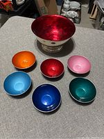 Top down view of large red enamel bowl and six smaller colorful enamel bowls arranged on gray surface.