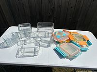Set of clear rectangular glass containers with matching plastic lids in blue, orange, and clear on white table outdoors, showing overall quantity and colors.