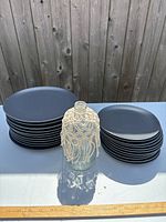 Eleven dark blue IKEA dinner plates, eleven matching side plates, and clear glass vase with cream macrame cover arranged outdoors on a grey surface with wooden fence backdrop.
