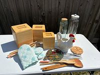 Overview of the kitchen assortment items on a white table outdoors including wooden storage bins, glass jars, ceramic chicken pitcher, oven mitts, wooden and metal utensils, and grill pins.