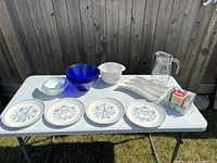 Full lot overview showing plates, bowls, pitcher, serving dishes, and salt & pepper set on white folding table outdoors.