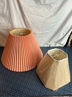 Photo showing two lamp shades on a black surface with a white backdrop; one large pink pleated antique style and one smaller off-white octagonal shade still wrapped in plastic.