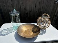 Three items displayed on a white surface: a clear glass jar with lid, a hammered brass-style bowl, and a round glass lantern wrapped in rope.