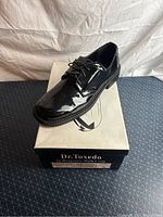 Side and top view of a single black patent leather men's tuxedo shoe on original box showing shiny finish and lace-up design.