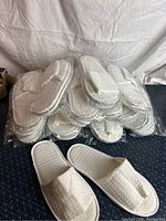 Stack of 15 pairs of white spa slippers, packaged in clear plastic, with one pair outside front and center to show detail.