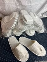 Photo showing multiple pairs of white waffle fabric spa slippers packaged in clear plastic and two pairs out of packaging.