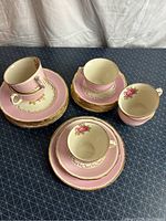 Full tea set arranged including six teacups, six saucers, six dessert plates and one cake plate with pink and gold design, floral pattern inside.