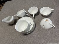 Top view of set of six white fine china teacups stacked on matching saucers, with one creamer cup placed to side, on a gray surface.