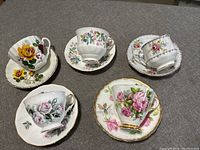 Five bone china floral teacups and saucers arranged on a grey surface with various floral patterns and gold trim