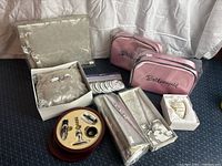 Full view of wedding gift set showing guest book, ring bearer pillow in box, two pink bridesmaid cosmetic bags, bachelorette pins, silver bar accessory set in case, silver purse clutch, and heart ornament.