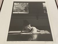 Black and white photograph depicting a partially nude woman draped with cloth posed on the floor in a sunlit room with window and outdoor trees visible.