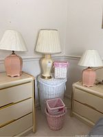 Three lamps and three wicker baskets arranged on and near two dressers against a wall. Two pink ceramic lamps with white fabric shades flank a cream ceramic lamp with a matching shade on top of the white wicker hamper. Small pink and white wicker baskets are positioned in front.