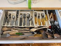 Top view of first drawer showing organized flatware with varied spoons, forks, knives, wooden spoons, and metal kitchen tools.
