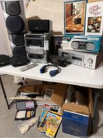 A collection on a folding table showing Aiwa stereo system, various board games, magazines, remote controls, boxed Sanyo portable radio cassette recorder, and other items.
