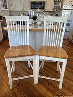 Two wooden bar stools with white painted frames and natural wood seats placed in a kitchen against a white cabinet and wood floor.