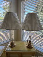 Two brass and glass table lamps on a yellow cabinet near windows, each with white pleated lampshades