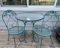 Front view of green metal bistro table with glass top flanked by two chairs on wooden deck in front of house siding