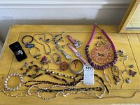 Full overhead view of the mixed lot of costume jewelry pieces on a wooden dresser including necklaces, bracelets, watches, earrings, rings, and a pair of costume jewelry pins.