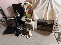 Wide shot showing multiple electronics including a large wide-format printer, CRT monitor, cords, telephones, and black camera on a table against a basement wall