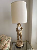 Front view of the figural lamp showing the boy figure holding a jug with a white cylindrical shade on a glass table.