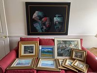 Wide shot showing all 10 framed artworks arranged on a couch including one large abstract piece and nine smaller framed landscape and cityscape paintings