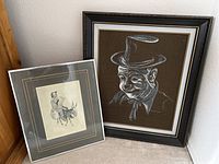 Two framed drawings resting against wall: one signature clown figure, another monochrome woman on donkey.