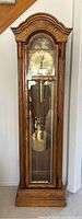 Full front view of tall wooden grandfather clock showing brass clock face, pendulum, weights, and glass door locked.