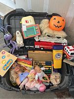 Plastic tub filled with assorted used toys including dolls, a toy fire truck, abacus, novelty items, and a Halloween pumpkin bucket.