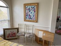 Six wooden TV trays with folding legs stacked next to two metal frame chairs with patterned cushions, and two framed floral paintings positioned on floor and wall.