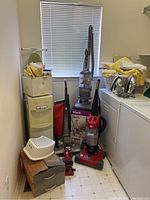 Wide view of the laundry room showing three vacuum cleaners arranged near the washing machine and dryer, water cooler, step stool, and folded laundry.