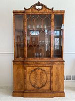Front view of vintage burl veneer wood hutch showing glass upper cabinet with decorative wooden overlays and carved top crest. Lower cabinet with large carved oval centerpiece and burled wood finish.