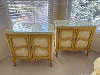 Pair of vintage French Provençal nightstands with yellow gold finish, sitting near window. Showing overall shape and condition with glass tops reflecting window light.