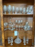Shelf of assorted vintage glassware including various stemmed glasses, small tumblers, and a faceted decanter. Glasses show various bowl shapes and stem styles. Hutch wood background visible.