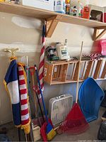 Photo showing wooden ladder, flags on poles, cleaning tools, blue sled, white box fan and shelves with various items.