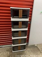 Front view of five stacked plastic storage bins with dark translucent front doors and brown handles against a red wall.