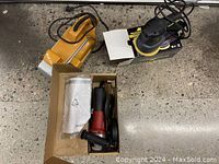 Lot showing three power tools: yellow B&D belt sander, black McCulloch random orbit sander on box, and red/black angle grinder in a box with accessories