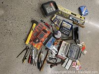 Assorted small hand tools and hardware arranged on a concrete floor, includes pliers, screwdrivers, socket set, hammer, levels, drill bits, and other accessories in original packaging or loose.