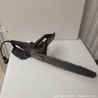 Side angle of black electric chainsaw on white surface showing entire length of blade and cord.