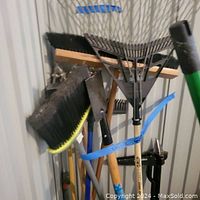 Photo showing garden tools including brooms, leaf rake, and ice chipper leaning against a wall secured with blue tape labeled Lot 2.
