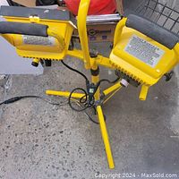 Halogen work lamp with two yellow light heads mounted on a yellow metal tripod stand on concrete floor.