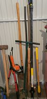 Photo showing a variety of long-handled garden tools leaning against a wall including a hedge trimmer, sledgehammer, and garden fork.