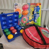 Complete lot showing bocce ball set with balls in blue case, Super Soaker blaster packaged, Squiggle pool toys packaged, and red round case bag for Rotorball.