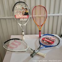 Four tennis rackets on table; includes two unused Wilson 23-inch rackets and two used rackets in orange-red and blue colors.